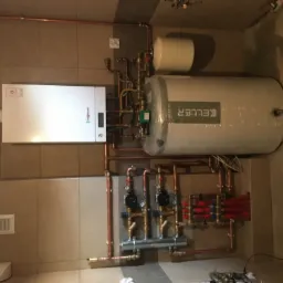 Nowa instalacja grzewcza z piecem gazowym Viessmann, zasobnikiem Keller i rozdzielaczem, rury miedziane, w folii ochronnej, na tle płytek ceramicznych.
