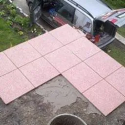 Układanie różowej kostki brukowej w kształcie litery L na podłożu z widoczną zaprawą, z otwartym bagażnikiem samochodu w tle.