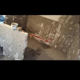 Surowy stan łazienki w trakcie prac: widoczne czerwone rury instalacji wodnej na ścianie z resztkami starej glazury, bloczki z betonu komórkowego oraz wiadro i packa murarska.