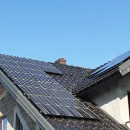 Panele słoneczne na dachu domu jednorodzinnego z brązową dachówką, widoczne fragmenty elewacji w trakcie wykończenia.