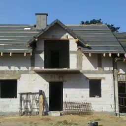 Surowy, dwukondygnacyjny budynek z białego pustaka w trakcie budowy dachu, z widocznymi łatami i wstępnym pokryciem, z drabina opartą o ścianę boczną i rusztowaniami przed budynkiem.