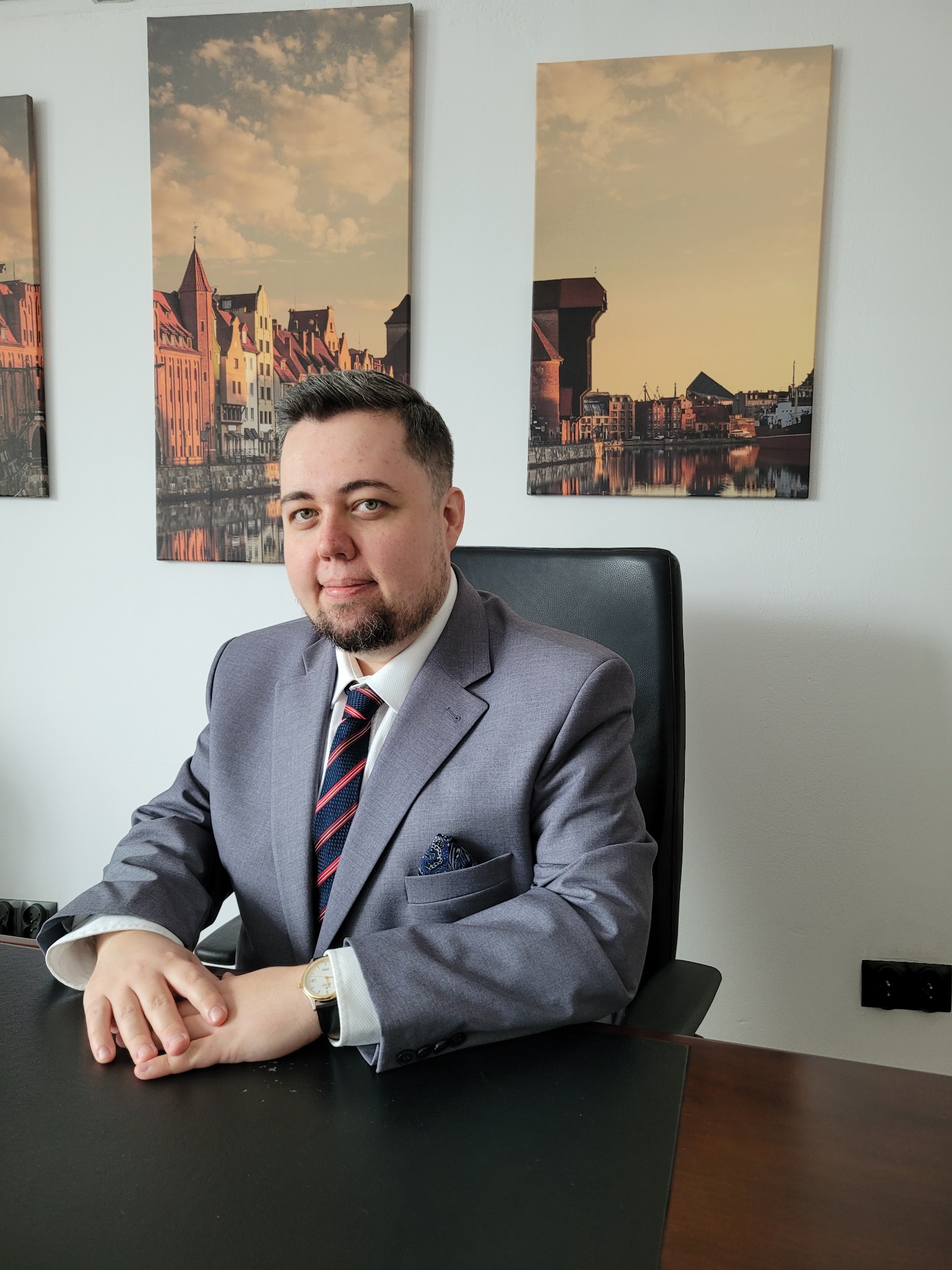 Mężczyzna w szarym garniturze siedzący za biurkiem, z obrazami Gdańska w tle.