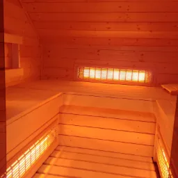 Wnętrze drewnianej sauny z ławkami i grzejnikiem na ścianie, oświetlone ciepłym, pomarańczowym światłem.