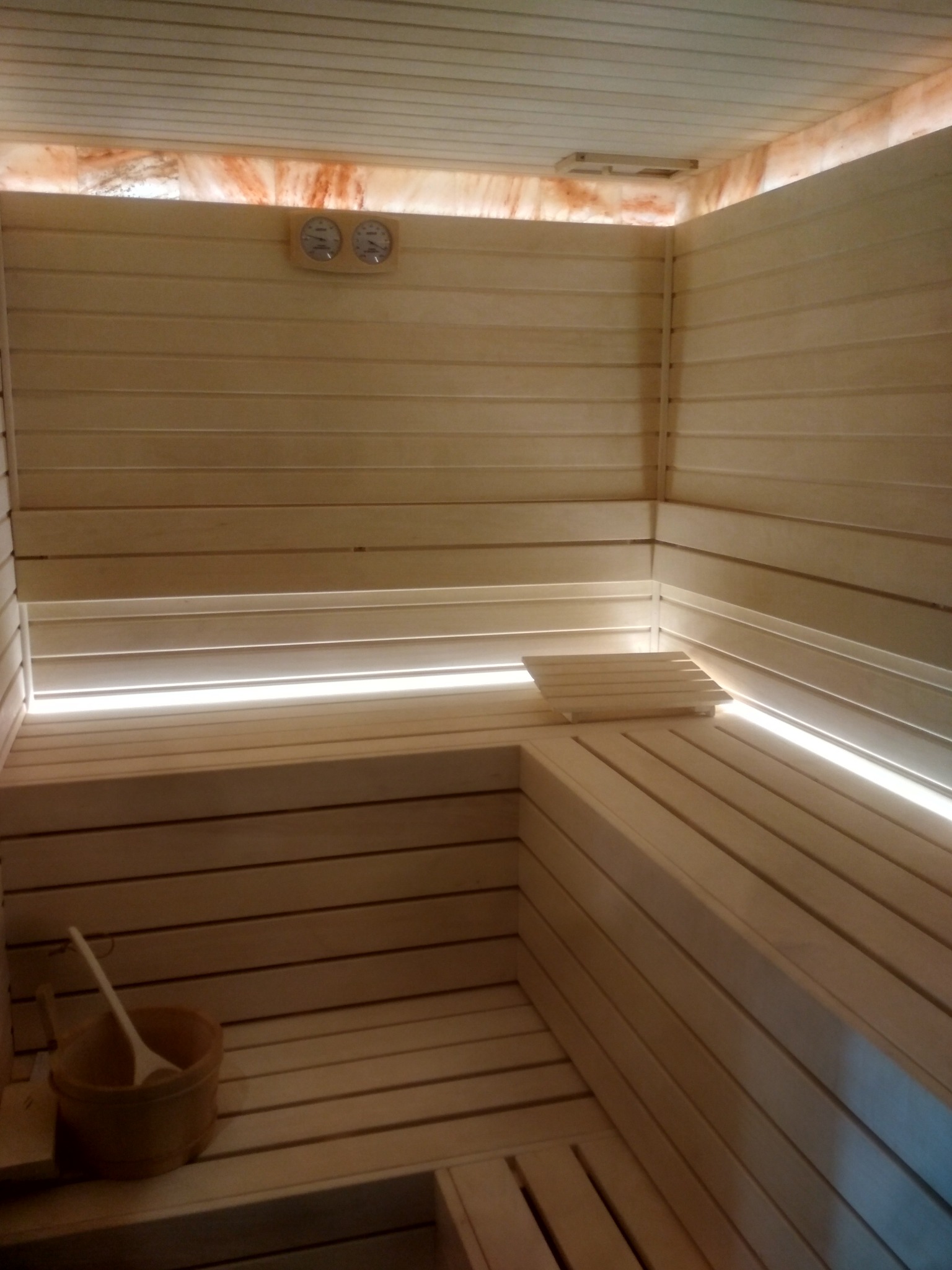 Wnętrze nowej, jasnej sauny z drewnianymi ławkami, oświetleniem LED pod siedziskami, drewnianym wiadrem z chochlą oraz termometrem i higrometrem na ścianie