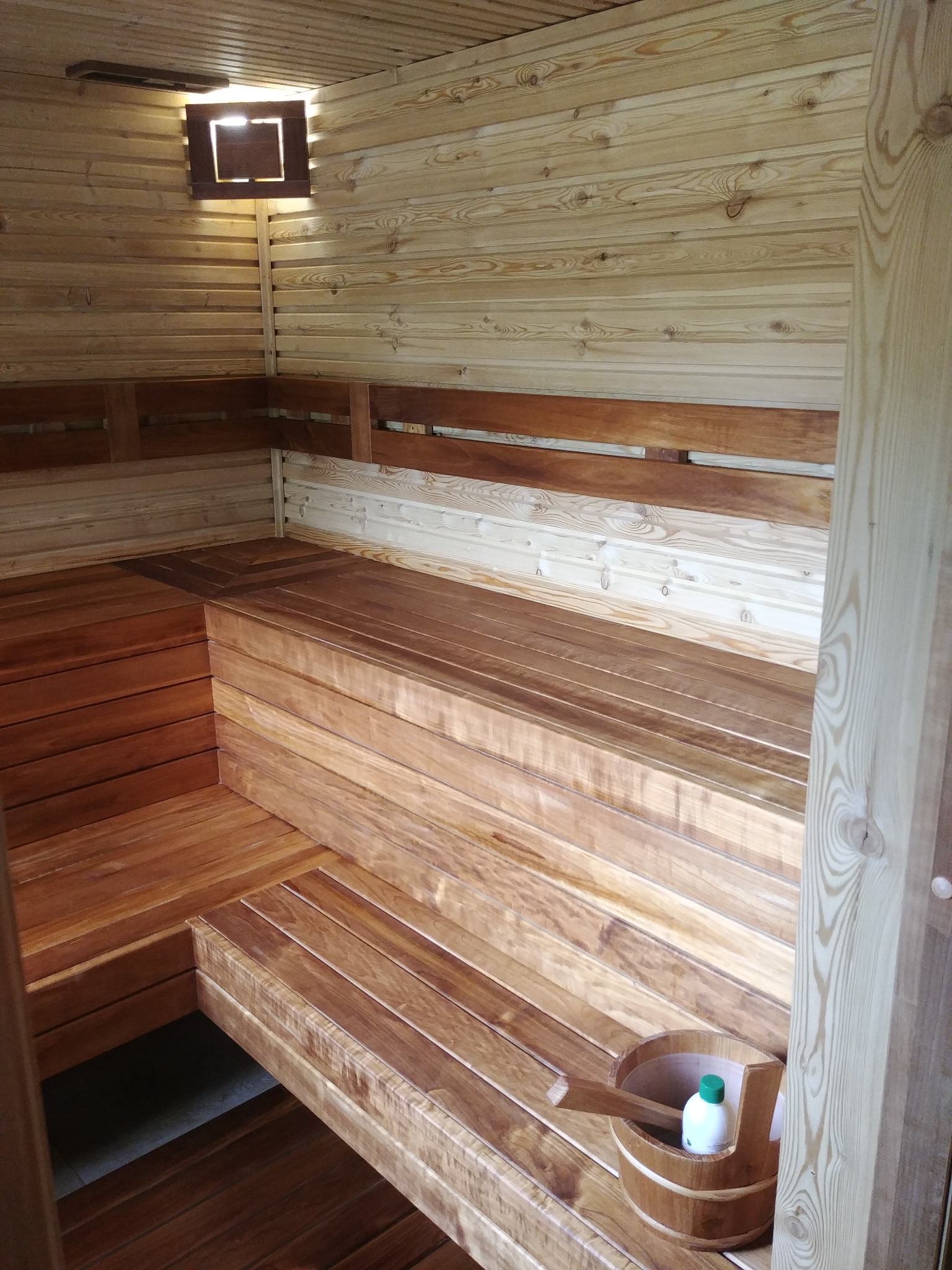 Wnętrze sauny wyłożone jasnym drewnem, z drewnianymi ławkami na różnych poziomach i oświetleniem w rogu, wiadro z akcesoriami na dolnej ławce.