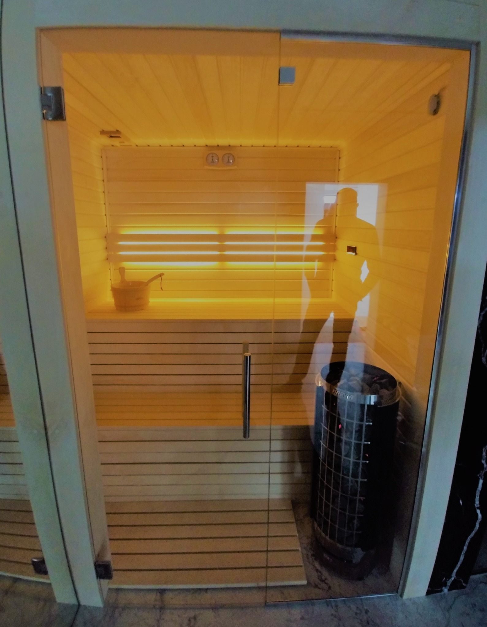 Nowoczesna sauna z przeszklonymi drzwiami, widoczne drewniane ławy, piec z kamieniami i odbicie sylwetki osoby na szkle.