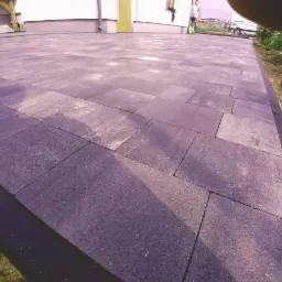 Ułożona szara kostka brukowa na podjeździe, widok z perspektywy bliskiej ziemi, z fragmentem osoby w tle i elementami ogrodu.