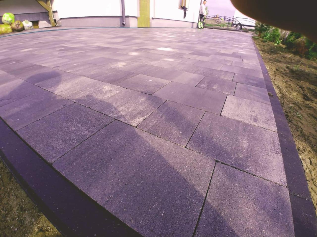 Ułożona szara kostka brukowa na podjeździe, widok z perspektywy bliskiej ziemi, z fragmentem osoby w tle i elementami ogrodu.