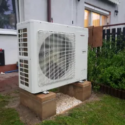 Biała zewnętrzna jednostka pompy ciepła Viessmann zainstalowana na betonowych blokach w Smardzowicach, otoczona zielenią, z widocznymi kroplami wody na obudowie.