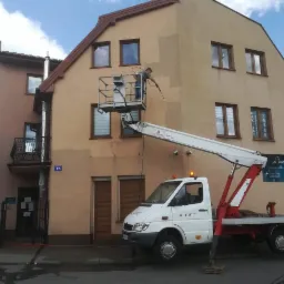 Pracownik na podnośniku koszowym aplikuje środek na elewację budynku, widoczne ślady czyszczenia i reklama salonu kosmetycznego na ścianie.