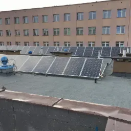 Widok z góry na rząd paneli słonecznych na płaskim dachu budynku z wieloma oknami; widoczne kominy wentylacyjne i niebieska instalacja wentylacyjna.