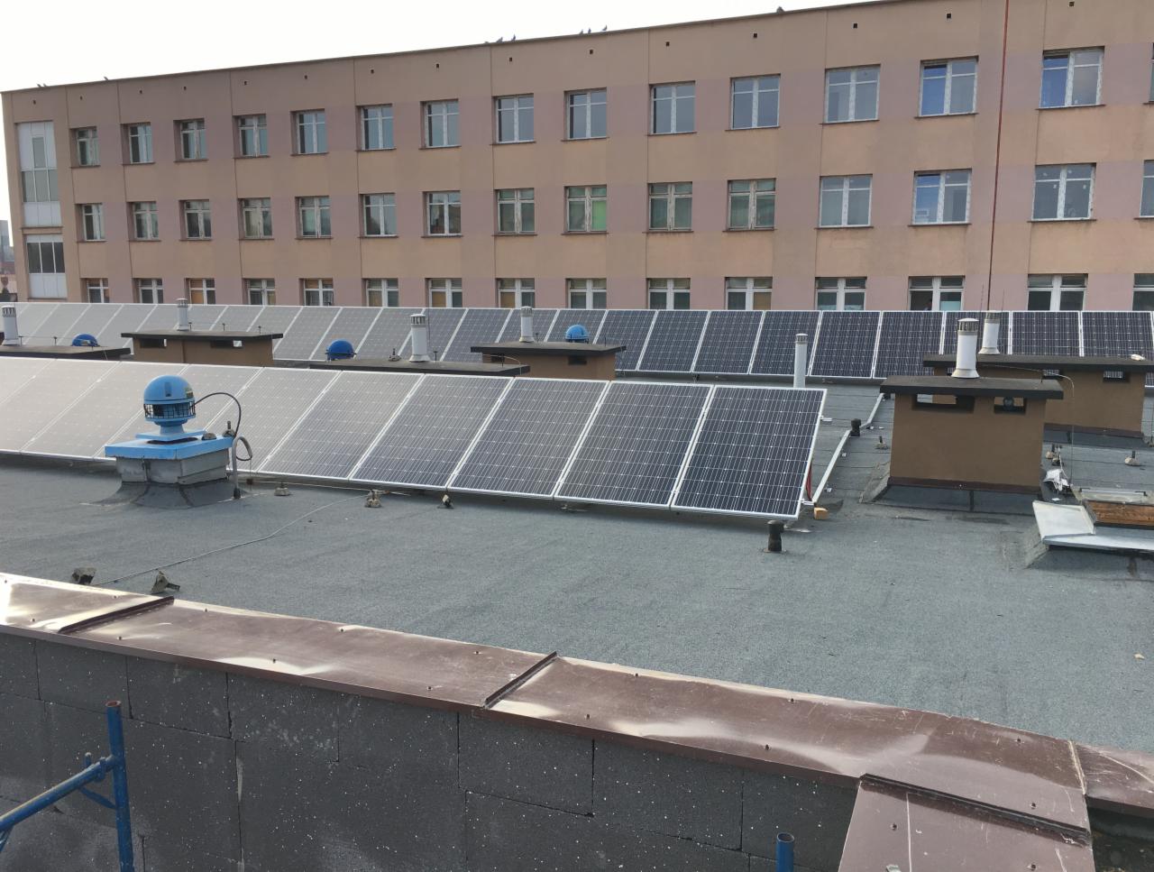 Widok z góry na rząd paneli słonecznych na płaskim dachu budynku z wieloma oknami; widoczne kominy wentylacyjne i niebieska instalacja wentylacyjna.