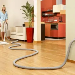 Kobieta odkurza drewnianą podłogę odkurzaczem centralnym; w tle widoczny salon z białymi meblami i kuchnia w odcieniach pomarańczy.