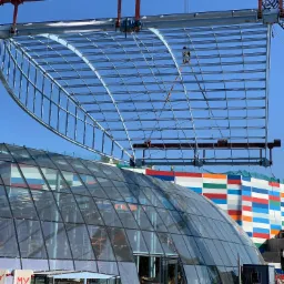 Montaż stalowej konstrukcji dachu z kratownic na tle przeszklonego budynku i kolorowej ściany, widoczny żuraw budowlany.