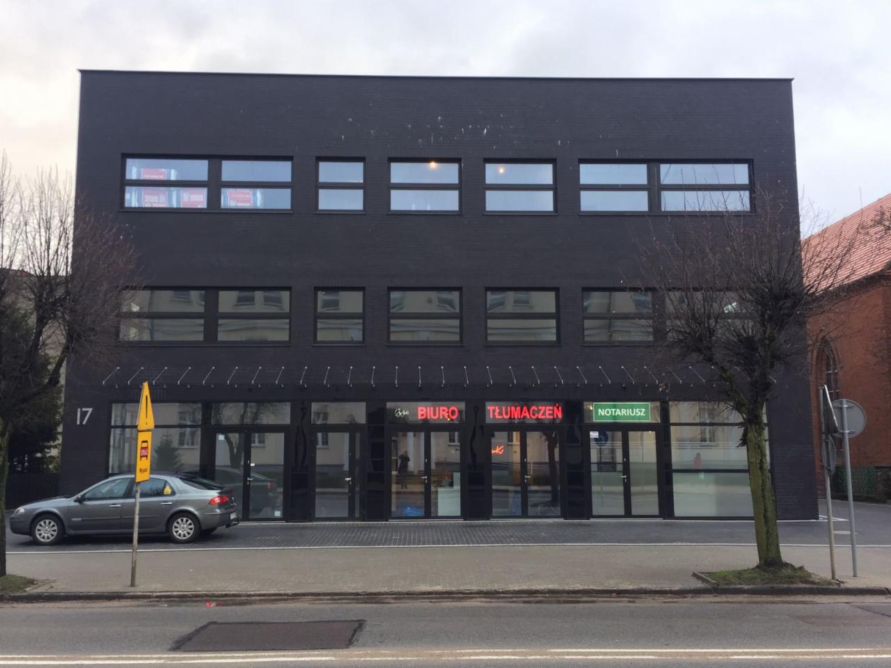 Fasada budynku z ciemnej cegły w Wejherowie, z neonowymi szyldami 'Biuro Tłumaczeń' i 'Notariusz' nad wejściami, widok z ulicy z zaparkowanym samochodem.