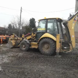 Żółta koparko-ładowarka CAT stoi na błotnistym terenie, z przodu widoczny pług do odśnieżania, na szybie naklejony numer telefonu.