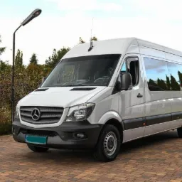 Biały bus Mercedes-Benz Sprinter z anteną na dachu, zaparkowany na brukowanym podjeździe przed zielenią.