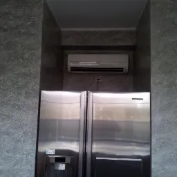 Klimatyzator ścienny zamontowany nad dużą, dwudrzwiową lodówką Samsung w niszy o szarych ścianach z efektem dekoracyjnym.