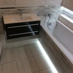 Nowoczesna łazienka z podświetlaną wanną, umywalką z szafką w kolorze czarnym i toaletą, wykończona płytkami imitującymi drewno.