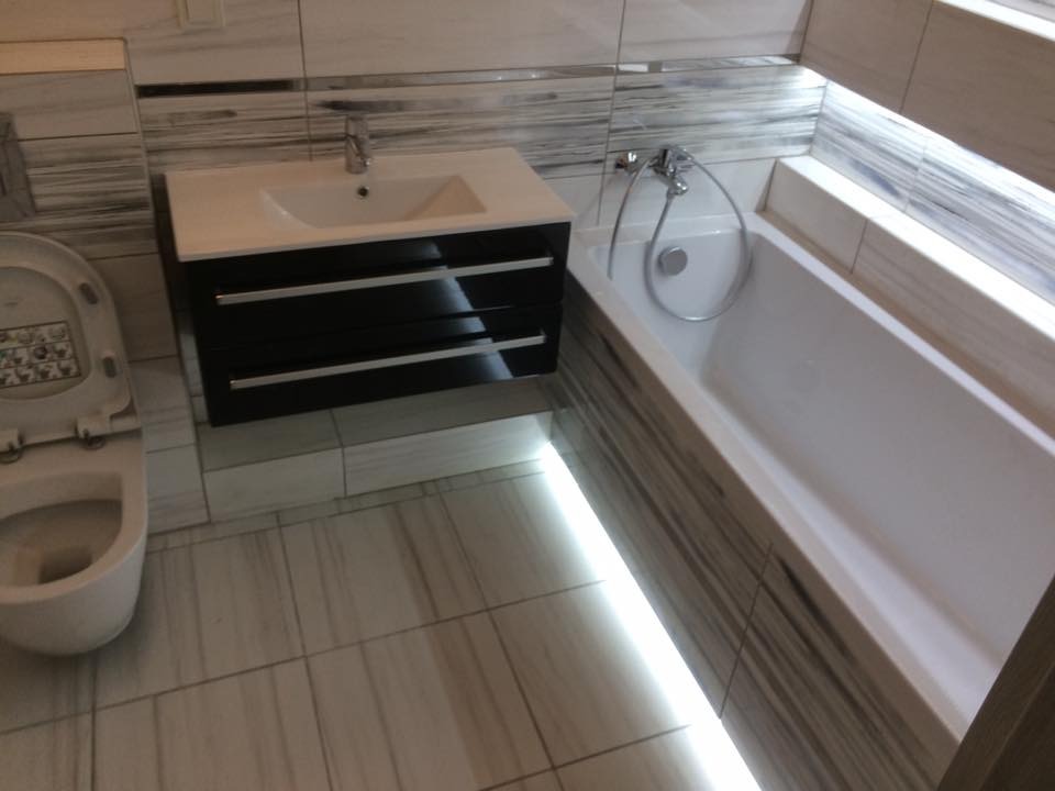 Nowoczesna łazienka z podświetlaną wanną, umywalką z szafką w kolorze czarnym i toaletą, wykończona płytkami imitującymi drewno.