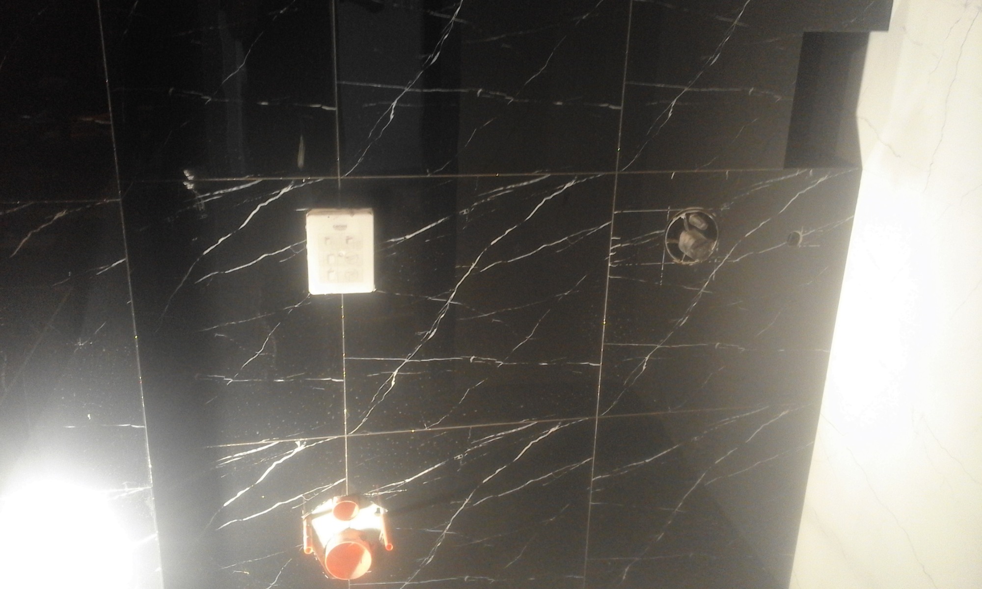 Czarne płytki imitujące marmur z białymi żyłami na ścianie łazienki, widoczna puszka elektryczna i otwór na wentylator, w dolnej części fragment zabudowy podtynkowy z rurami.