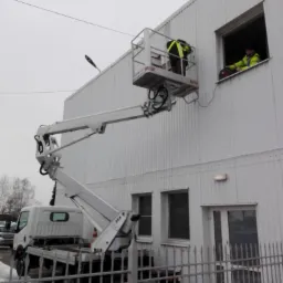 Pracownicy w żółtych kamizelkach pracują na podnośniku koszowym przy oknie budynku przemysłowego z blachy trapezowej, widoczne ogrodzenie metalowe i śnieg.