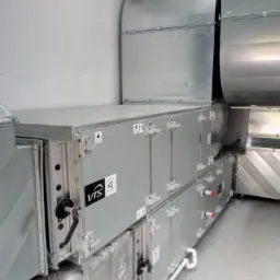 Szara centrala wentylacyjna VTS z widocznymi klapami rewizyjnymi i rurami, zainstalowana w pomieszczeniu technicznym.