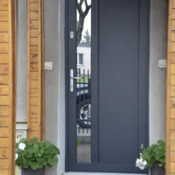 Szare drzwi wejściowe z wąskim przeszkleniem, odbijającym fragment drzewa i samochodu, zamontowane w betonowej ścianie, z drewnianą ramą werandy po bokach i donicami z kwiatami po obu stronach...