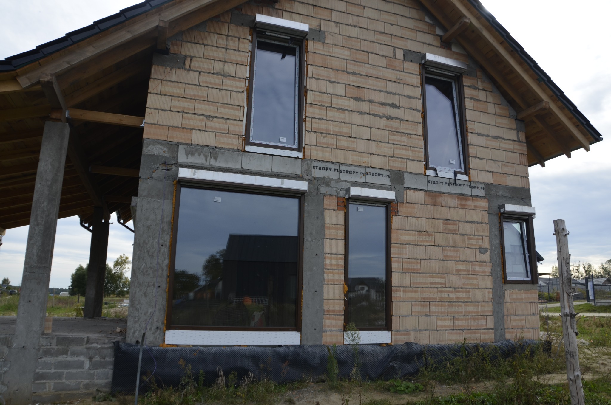Surowy stan budynku jednorodzinnego w trakcie montażu okien z brązowymi ramami, widoczne białe profile montażowe i niezakończona elewacja z cegły ceramicznej.