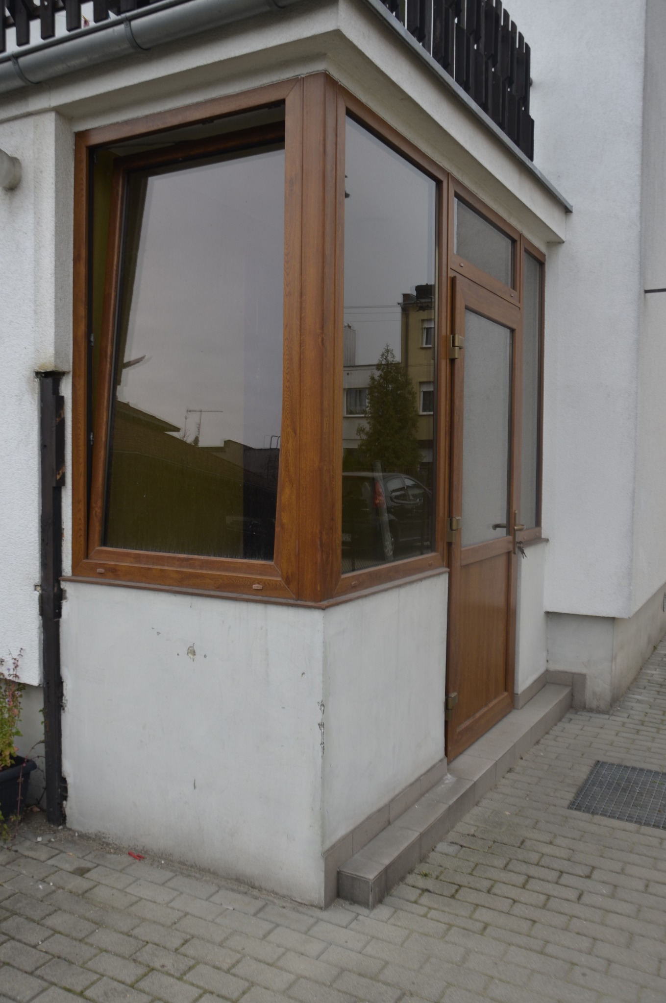 Zewnętrzne wejście do budynku z drewnianą stolarką okienną i drzwiową, częściowo otwarte okno uchylne, szara kostka brukowa i biały tynk.
