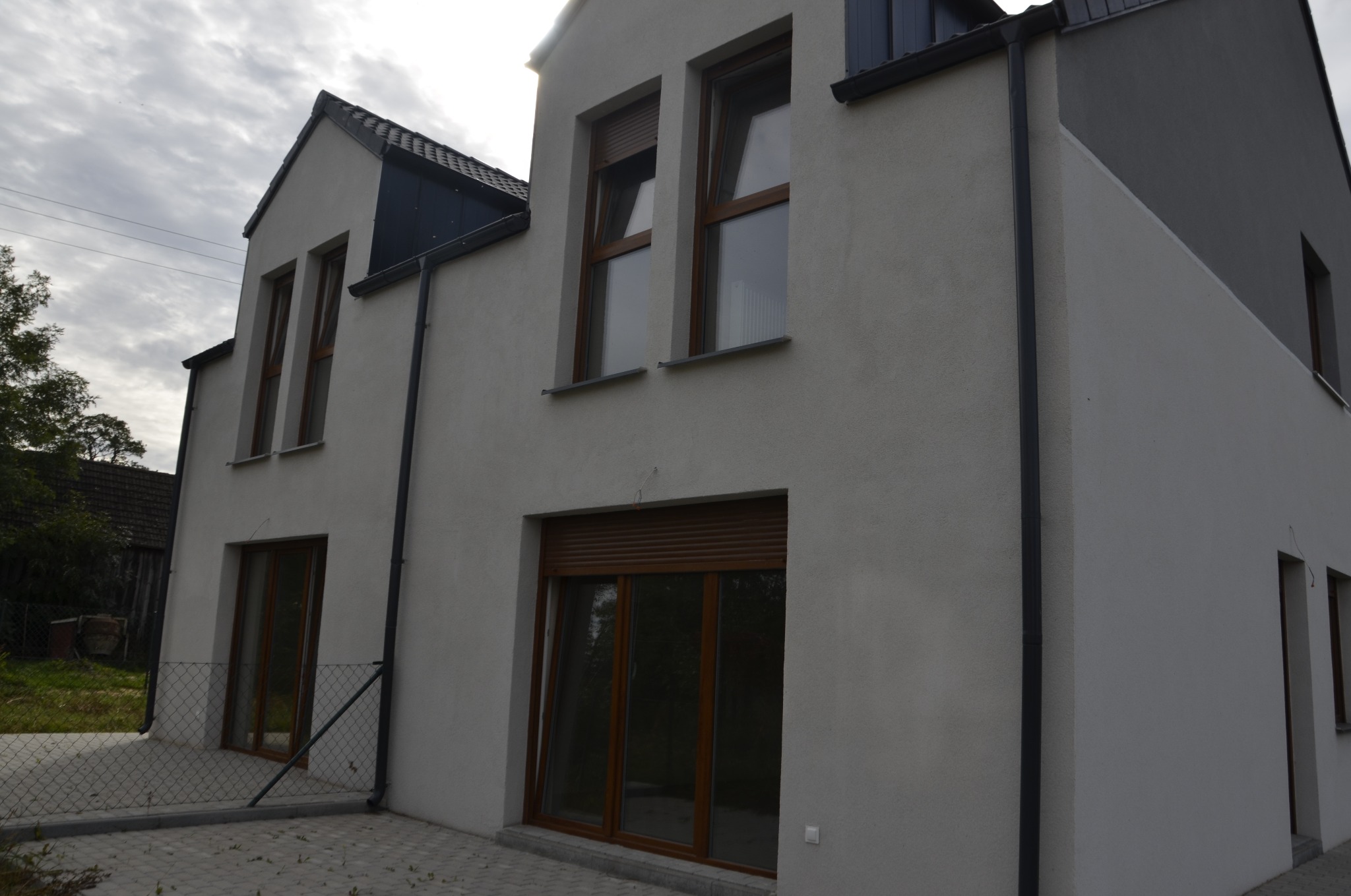 Nowoczesny dom jednorodzinny z brązowymi oknami i roletami zewnętrznymi, widok z boku na elewację wykończoną w odcieniach szarości i bieli, z widoczną metalową siatką ogrodzeniową w tle.