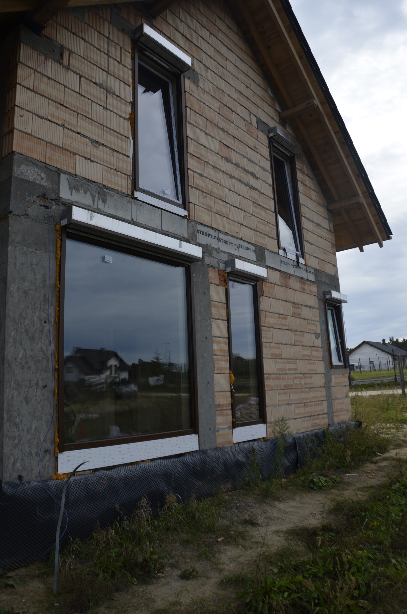 Surowy stan elewacji domu jednorodzinnego z zamontowanymi oknami z brązowymi ramami i roletami zewnętrznymi w kolorze białym, widoczne detale montażu i niedokończone ocieplenie.