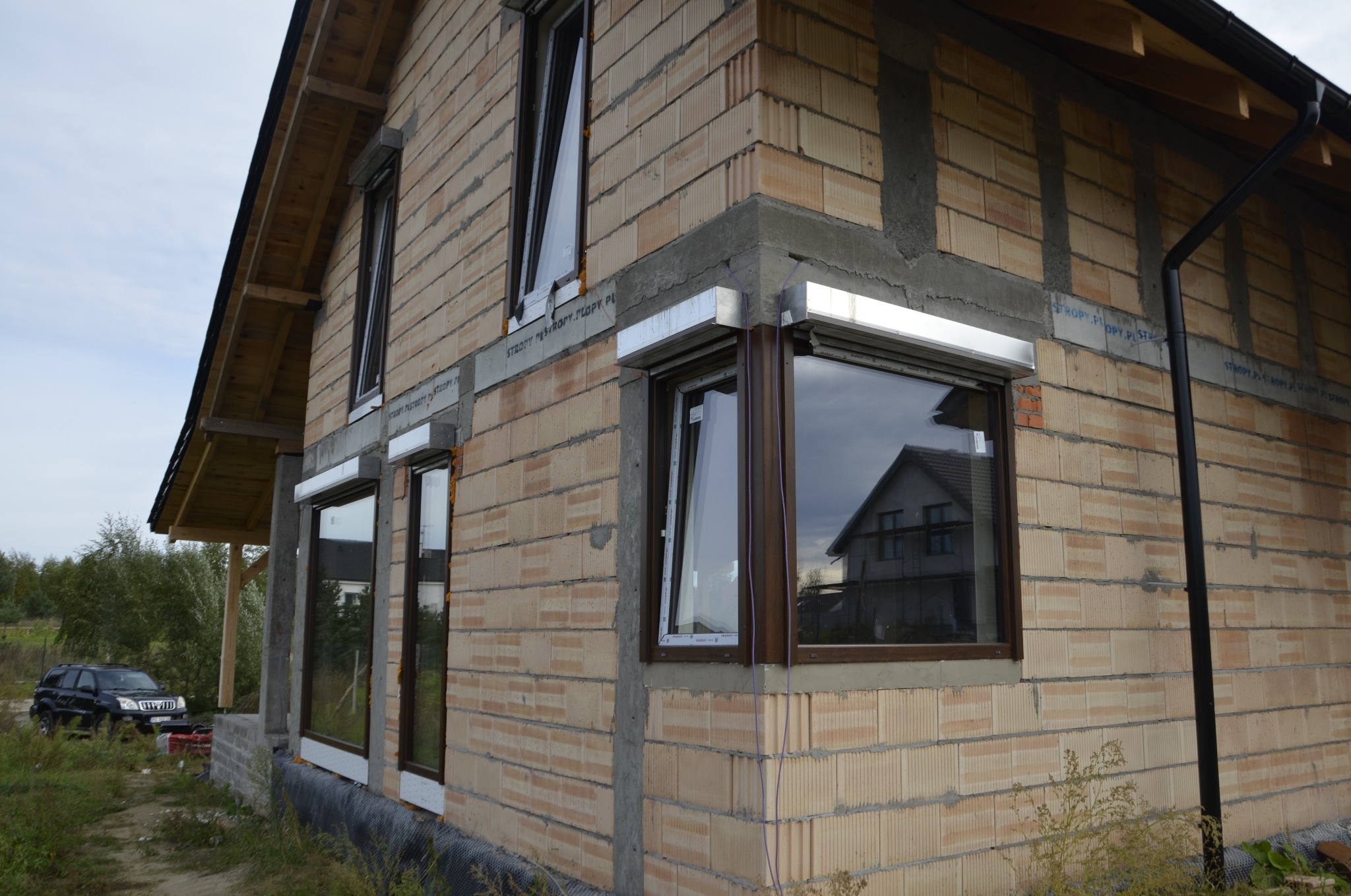 Surowy stan elewacji z zamontowanymi oknami z brązowymi ramami i roletami zewnętrznymi, widoczny fragment dachu i rynna, w tle dom sąsiada.