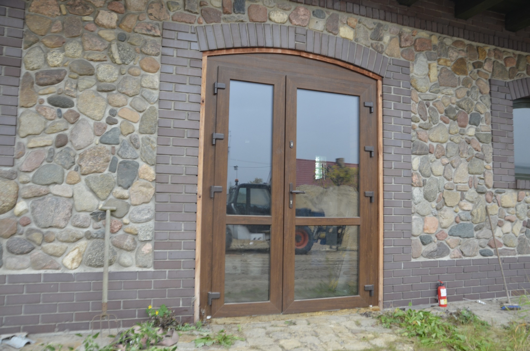 Dwuskrzydłowe drzwi z brązowymi ramami i szklanymi panelami, wkomponowane w elewację z kamienia i cegły, z widocznym odbiciem traktora.