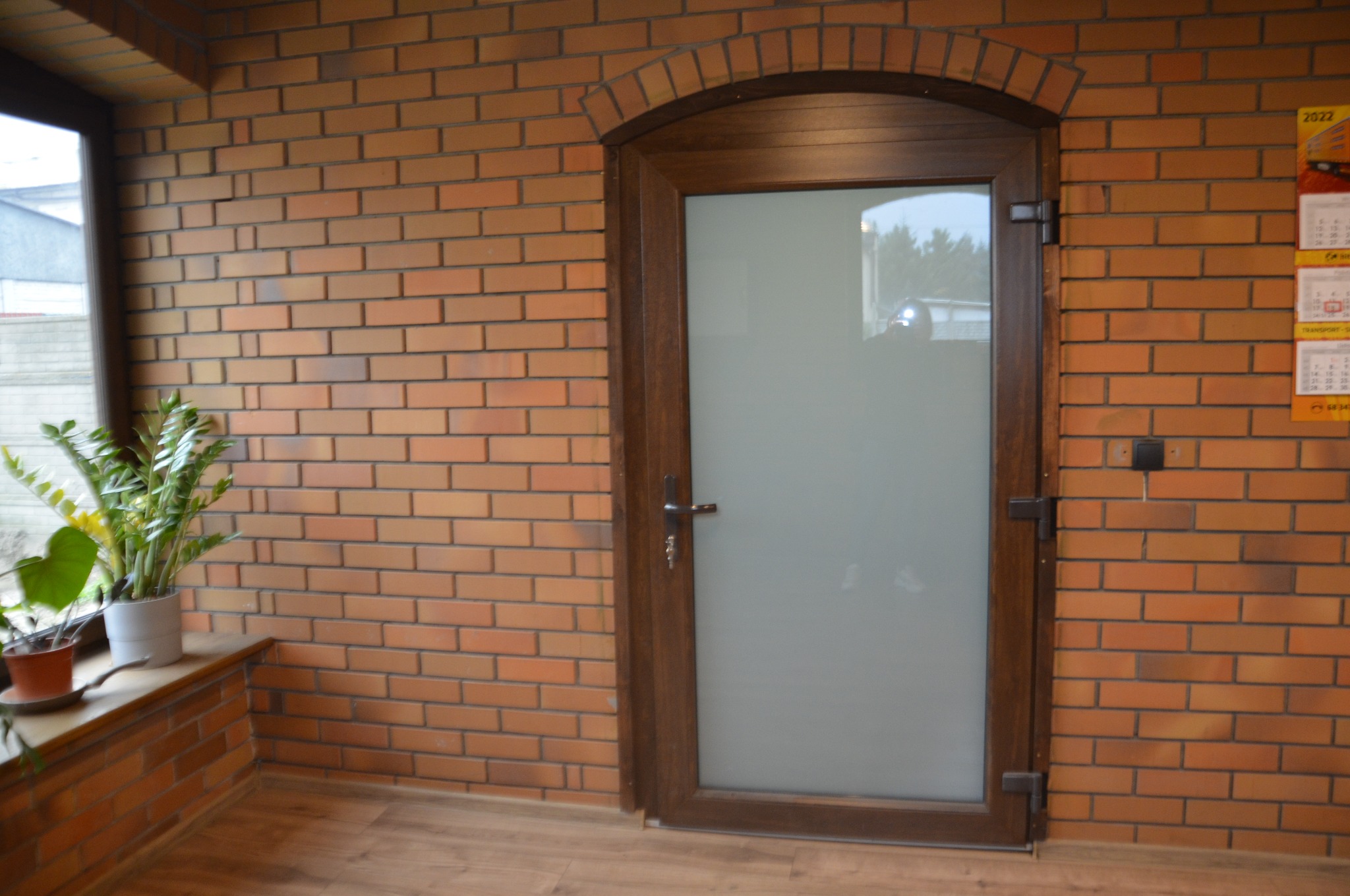 Wnętrze pomieszczenia z ceglaną ścianą, drewnianą podłogą i przeszklonymi drzwiami z drewnianą ramą o łukowatym kształcie, obok okna z roślinami doniczkowymi.