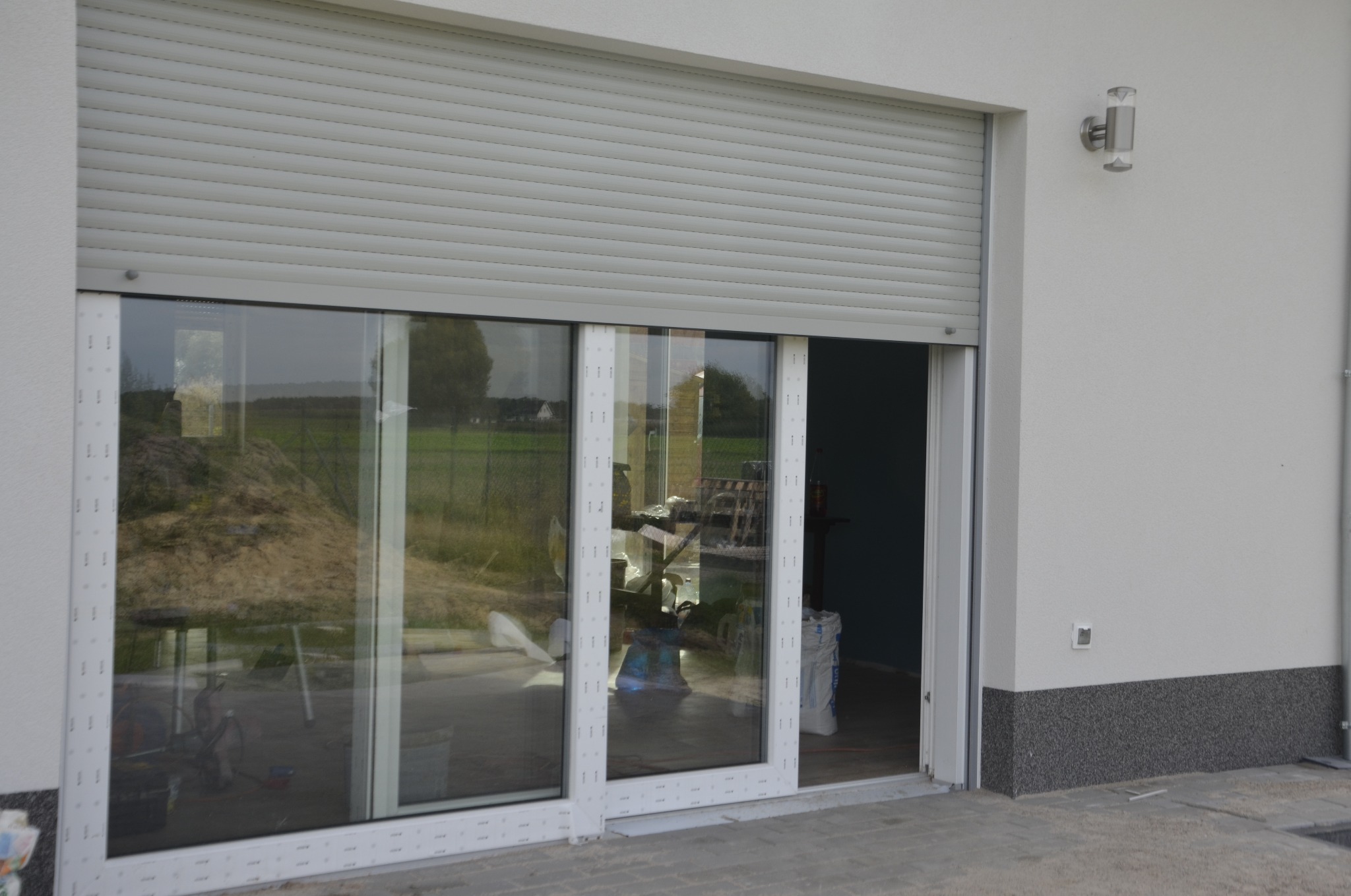 Trzyczęściowe przeszklone drzwi przesuwne z częściowo opuszczoną szarą roletą zewnętrzną, widok na plac budowy przez szybę, zewnętrzna lampa ścienna.