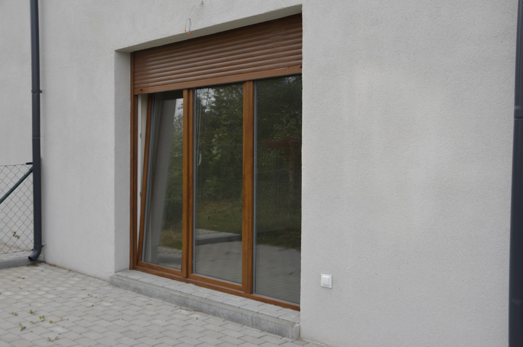 Brązowe okno z roletami zewnętrznymi, jedno skrzydło uchylone, widok z zewnątrz na elewację budynku.