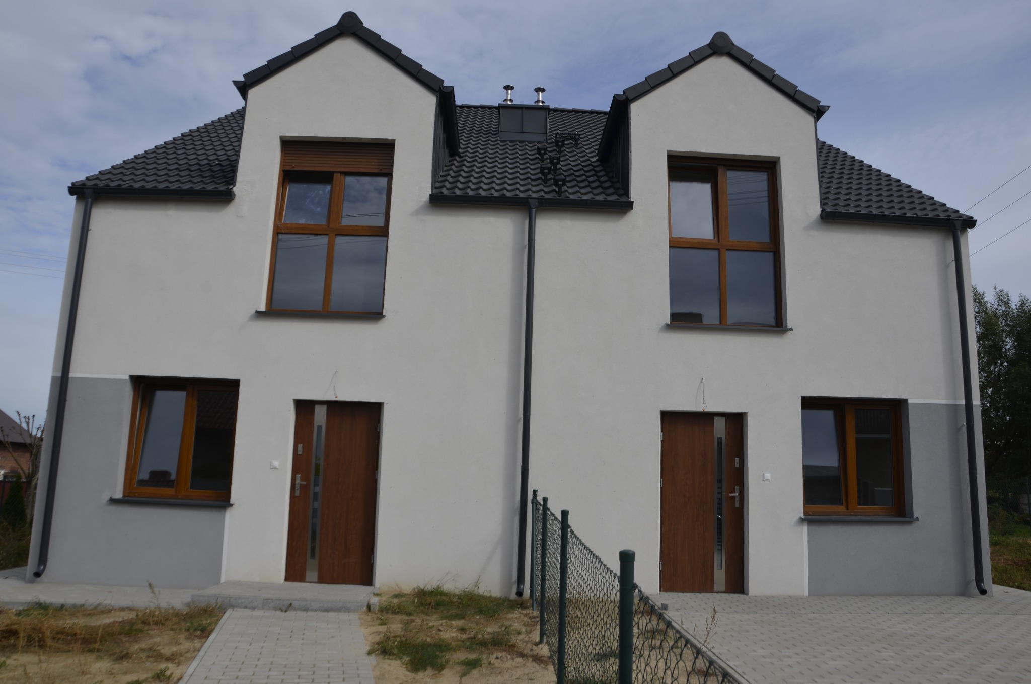 Nowo wybudowany bliźniak z brązowymi oknami PCV i drzwiami wejściowymi, widok z zewnątrz na elewację w odcieniach bieli i szarości, z czarnym dachem i rynnami.