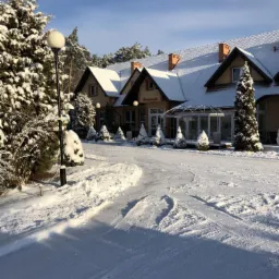 Zimowy krajobraz: budynek restauracji z przeszkloną werandą, pokryty śniegiem, otoczony ośnieżonymi drzewami i ścieżką z wyraźnymi śladami