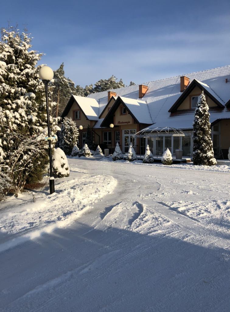 Zimowy krajobraz: budynek restauracji z przeszkloną werandą, pokryty śniegiem, otoczony ośnieżonymi drzewami i ścieżką z wyraźnymi śladami