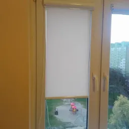 Biała, częściowo zwinięta roleta w oknie PCV z widokiem na plac zabaw.
