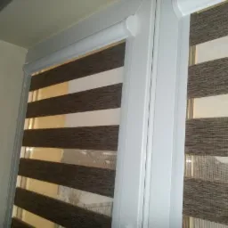 Fragment okna z zamontowaną, brązowo-białą, poziomą przesłoną okienną typu dzień i noc w białej ramie, widoczne z bliska.