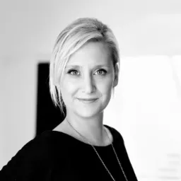 Portret kobiety z krótkimi blond włosami, ubranej w czarną bluzkę i srebrny łańcuszek, uśmiechającej się do kamery na jasnym tle.