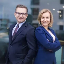 Portret dwójki uśmiechniętych profesjonalistów w garniturach, stojących plecami do siebie na tle szklanej fasady budynku z widocznymi odbiciami.