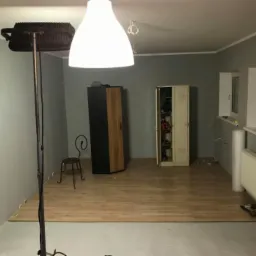 Pokój w trakcie remontu: częściowo ułożone panele podłogowe, widoczne narzędzia, grzejnik, szafa, metalowa szafka z otwartymi drzwiami, lampa podłogowa z reflektorem i wisząca lampa sufitowa.