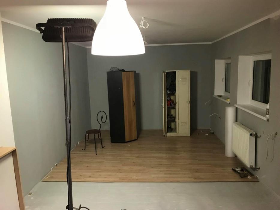 Pokój w trakcie remontu: częściowo ułożone panele podłogowe, widoczne narzędzia, grzejnik, szafa, metalowa szafka z otwartymi drzwiami, lampa podłogowa z reflektorem i wisząca lampa sufitowa.