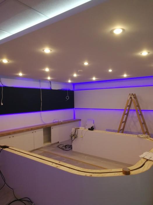 Niedokończona zabudowa z płyty karton gips z oświetleniem LED, widoczne kable, drabina i meble w tle.