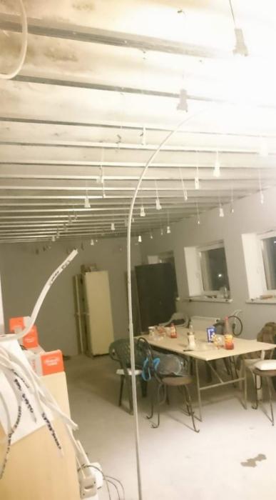 Widok wnętrza w trakcie prac wykończeniowych: metalowa konstrukcja sufitu podwieszanego, zwisające kable oświetleniowe, stół z resztkami jedzenia i napojów, narzędzia i materiały budowlane w tle.