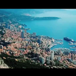 Panoramiczny widok z góry na Monako: gęsta zabudowa miejska z wieżowcami, port jachtowy z zacumowanym dużym wycieczkowcem, błękitne morze i zalesione góry w tle.