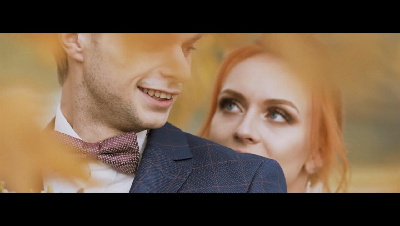 Uśmiechnięty pan młody w granatowej marynarce w kratę i bordowej muszce, spoglądający w bok, zza jego ramienia patrzy panna młoda z rudymi włosami i niebieskimi oczami.